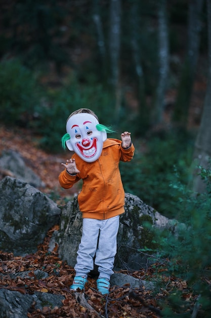Ein Kind mit Clownsmaske steht in einem Wald.