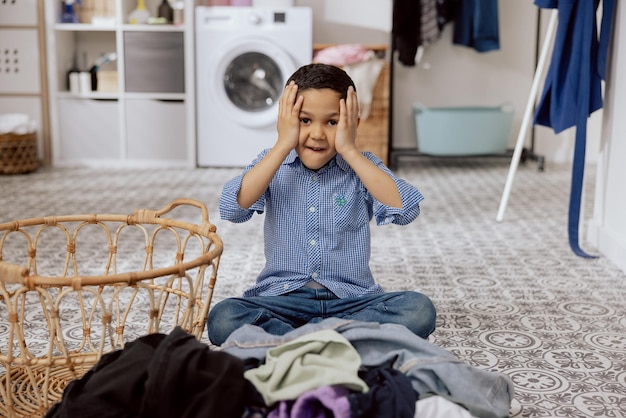 Ein Kind mit dunklen Haaren sitzt auf dem Boden der Waschküche im Badezimmer, ein verängstigter Junge, der sich den Kopf umklammert, macht Hausarbeit und sortiert müde eine riesige Menge an Kleidung