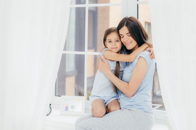 Ein Kind mit einem angenehmen Aussehen umarmt seine Mutter, drückt Liebe und ein gutes Gefühl oder eine gute Einstellung aus, setzt sich auf die Fensterbank und genießt die häusliche Atmosphäre