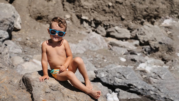 Ein Kind mit Sonnenbrille sitzt und sonnt sich auf großen Felsbrocken am Strand in der Nähe des Meeres