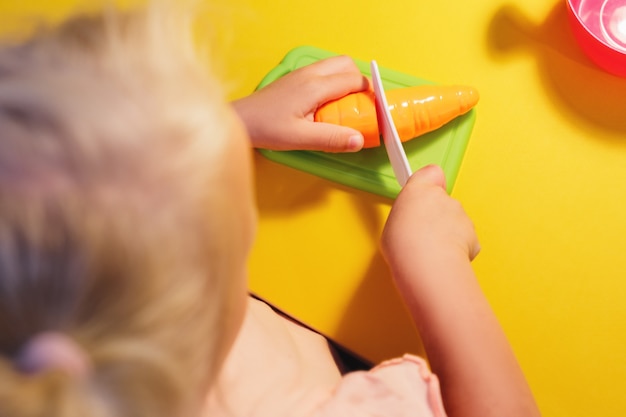 Ein Kind schneidet mit einem Spielzeugmesser eine Spielzeugkarotte auf gelbem Grund