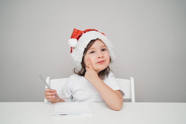 Ein Kind schreibt einen Brief an den Weihnachtsmann Ein süßes Mädchen in einer Weihnachtsmannmütze schreibt eine Todo-Liste Platz für...