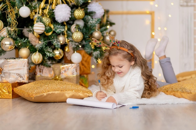 Ein Kind schreibt unter einem goldgeschmückten Weihnachtsbaum einen Brief für den Weihnachtsmann