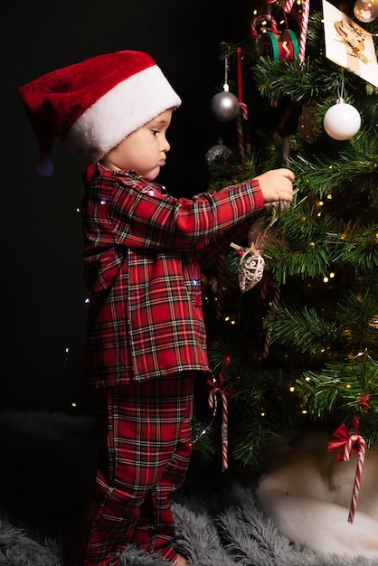 Ein Kinderjunge in Weihnachtsmütze und Pyjama hängt ein Spielzeug an den Weihnachtsbaum.