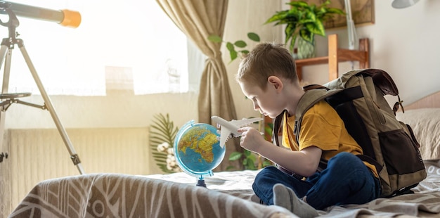 Ein Kinderjunge mit Rucksack spielt mit einem Spielzeugflugzeug und einem Erdball, der auf eine Reise in Richtung Abenteuer geht