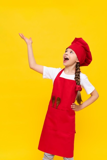 Ein Kinderkoch hält ein Tablett mit Ihrem Gericht über dem Kopf Ein kleines Mädchen in einer roten Kochmütze und einer roten Schürze auf gelbem, isoliertem Hintergrund