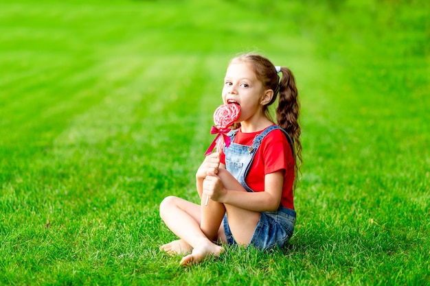 Ein Kindermädchen im Sommer auf dem Rasen mit einem großen Lutscher auf dem grünen Gras hat Spaß und freut sich, Platz für Text zu lecken