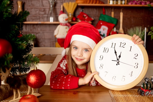 Ein Kindermädchen in einer Weihnachtsmütze mit einer großen Uhr in einer dunklen Küche an einem Weihnachtsbaum mit roten Kugeln freut sich und lächelt, das Konzept von Neujahr und Weihnachten