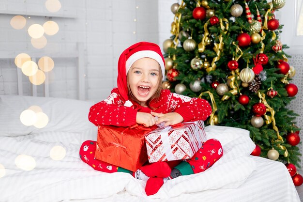 Ein Kindermädchen mit Geschenken am Weihnachtsbaum in rotem Pullover und Weihnachtsmannmütze an Silvester oder Weihnachten zu Hause auf einem weißen Bett schreit vor Glück