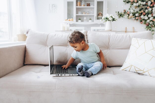 Ein kleiner afroamerikanischer dunkelhäutiger Junge sitzt mit einem Laptop auf einem Sofa