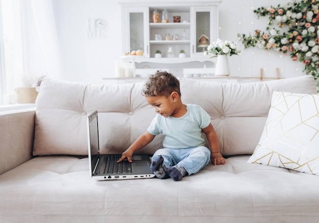 Ein kleiner afroamerikanischer dunkelhäutiger Junge sitzt mit einem Laptop auf einem Sofa