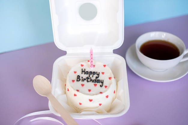 Ein kleiner Bento-Kuchen in Öko-Verpackung mit einem Holzlöffel und einer Kerze neben einer Tasse Tee auf einem violetten Hintergrund