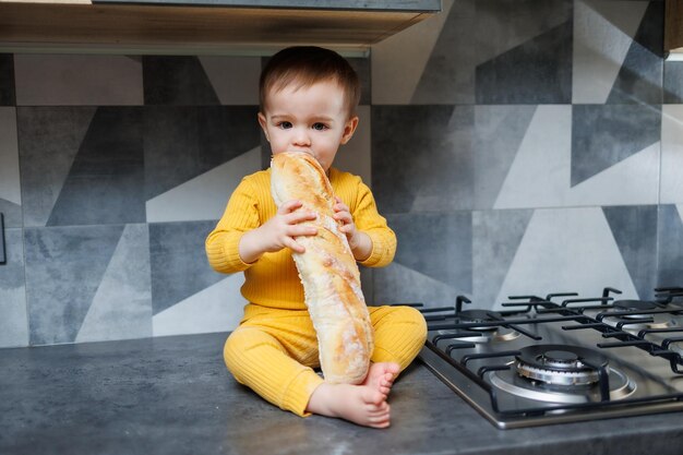 Ein kleiner einjähriger Junge sitzt in der Küche und isst in der Küche ein langes Brot oder Baguette