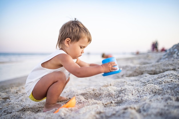 Ein kleiner fröhlicher, enthusiastischer Junge spielt mit seinen Spielsachen an einem Sandstrand, baut Perlen und Türmchen und lächelt jemanden hinter den Kulissen in einem hellen, sonnigen Sommerurlaub an