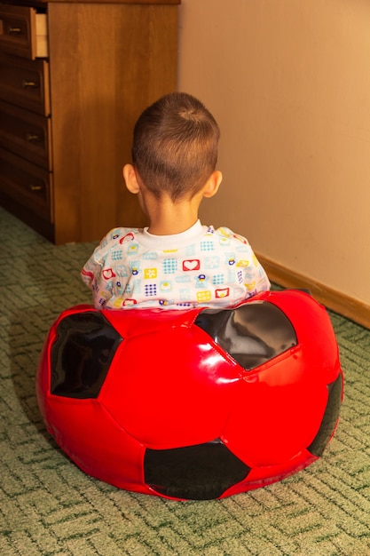 Ein kleiner gequälter Junge sitzt mit dem Gesicht zur Wand in einem weichen Sitzsack für Kinder in Form eines roten Fußballballs auf dem Boden