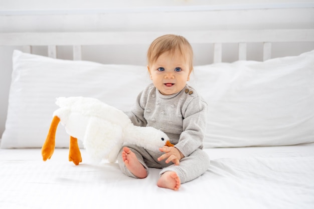 Ein kleiner Junge, ein blondes Kind mit blauen Augen in einem grauen Anzug, sitzt zu Hause im Schlafzimmer auf einem Bett mit weißer Bettwäsche und hält ein weiches Gänsespielzeug in den Händen