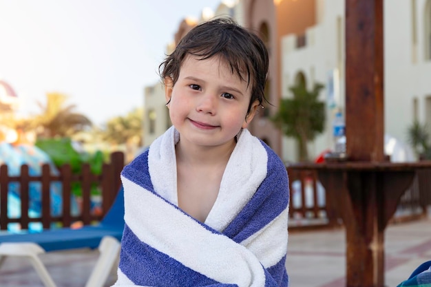 Ein kleiner Junge, eingewickelt in ein gestreiftes Handtuch, nachdem er in den Sommerferien mit Kindern in Luxushotels in einem Pool geschwommen war