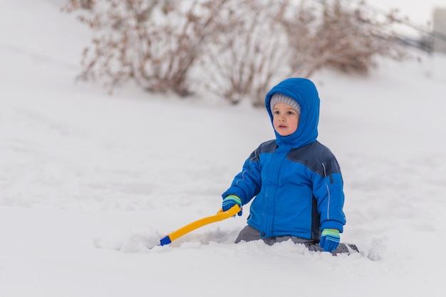 Ein kleiner Junge gräbt im Winter Schnee mit einer Schaufel.