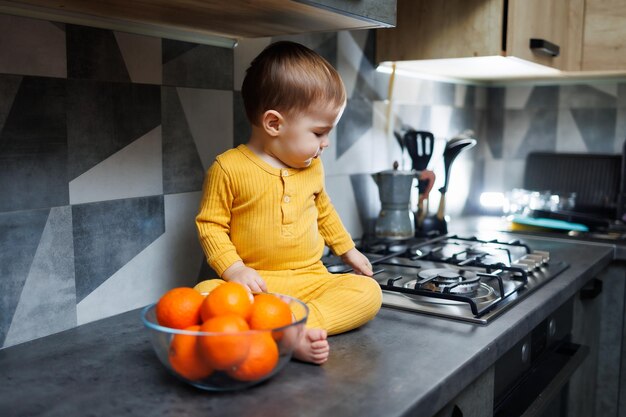 Ein kleiner Junge im Alter von 1 Jahr in gelber Kleidung sitzt in der Küche auf dem Tisch mit einem Teller mit orangefarbenen Mandarinen Porträt eines süßen einjährigen Jungen und süßer Zitrusfrüchte
