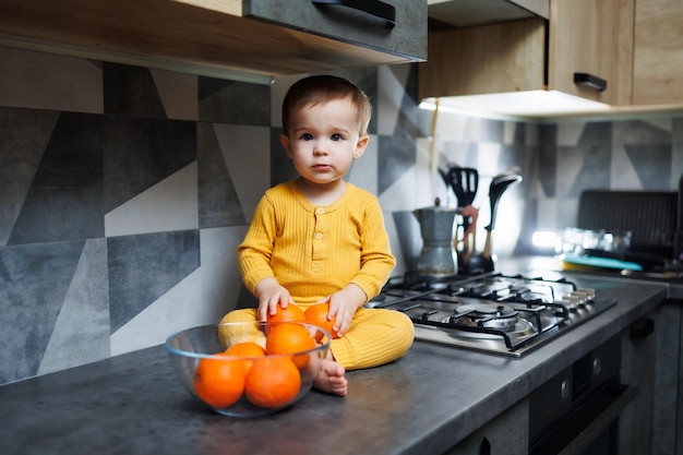 Ein kleiner Junge im Alter von 1 Jahr in gelber Kleidung sitzt in der Küche auf dem Tisch mit einem Teller mit orangefarbenen Mandarinen Porträt eines süßen einjährigen Jungen und süßer Zitrusfrüchte