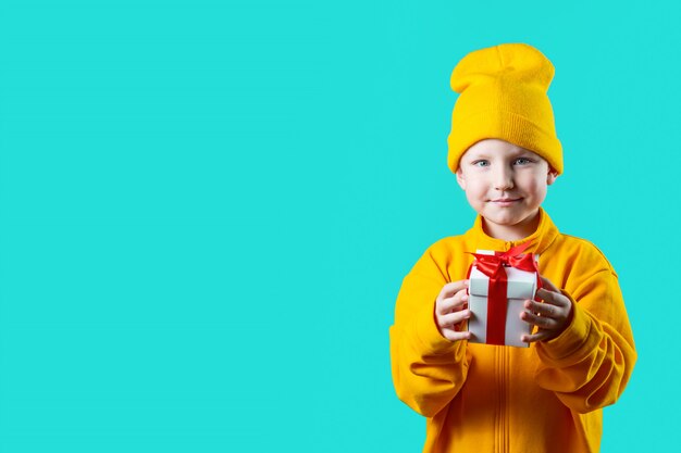 Ein kleiner Junge im gelben Hut und in der Jacke, die ein Geschenk auf einem tadellosen Hintergrund hält