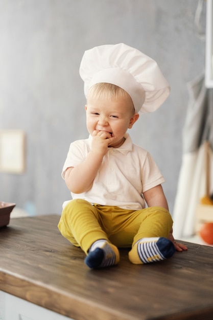 ein kleiner Junge im Hut des Chefs in der Küche, die auf dem Tisch sitzt und isst