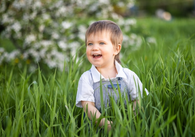 Ein kleiner Junge im Jeansoverall mit ausdrucksstarken blauen Augen. Springen und herumalbern im hohen grünen Gras vor dem Hintergrund eines großen grünen Busches und eines blühenden Hausgartens.