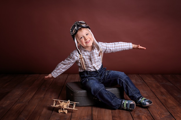 Ein kleiner Junge im Pilotenhelm spielt mit einem Holzflugzeug. Junge in einem karierten Hemd und Hosenträgern.