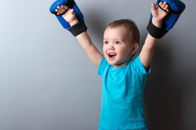 Ein kleiner Junge in Boxhandschuhen und einem blauen T-Shirt auf grauem HintergrundAusdruck von EmotionenKopierraum