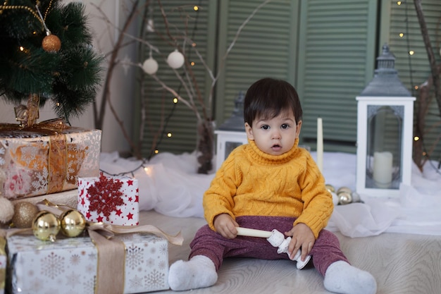 Ein kleiner Junge in einem gelben Pullover sitzt auf dem Boden und spielt mit einer Kerze in der Nähe des Weihnachtsbaums und einem festlichen Interieur.