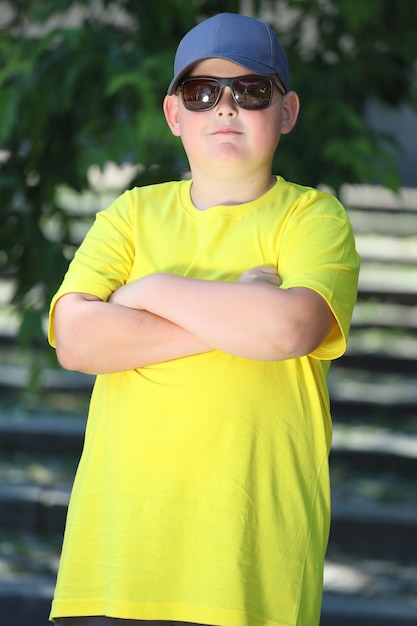 Ein kleiner Junge in einem gelben T-Shirt und Mütze und Sonnenbrille steht in der Natur. Foto in hoher Qualität