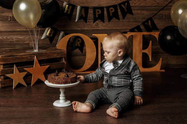 Ein kleiner Junge in einem grauen Anzug feiert seinen ersten Geburtstag und bricht einen Kuchen auf braunem Hintergrund mit Dekor