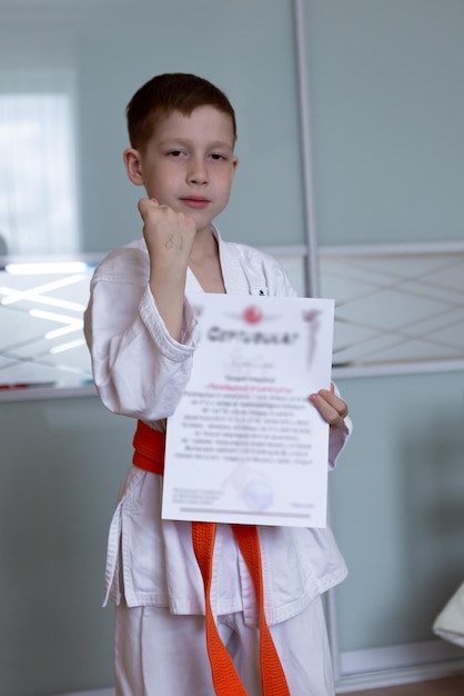 Ein kleiner Junge in einem Kimono mit orangefarbenem Gürtel steht mit einem Diplom in den Händen auf hellem Hintergrund zu Hause