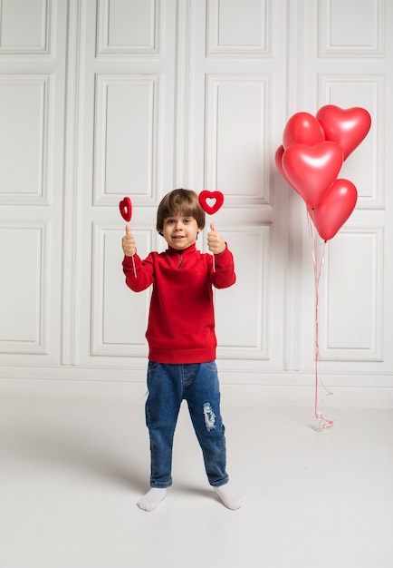 Ein kleiner Junge in einem Pullover und Jeans hält Herzen auf Stick auf Weiß mit Luftballons