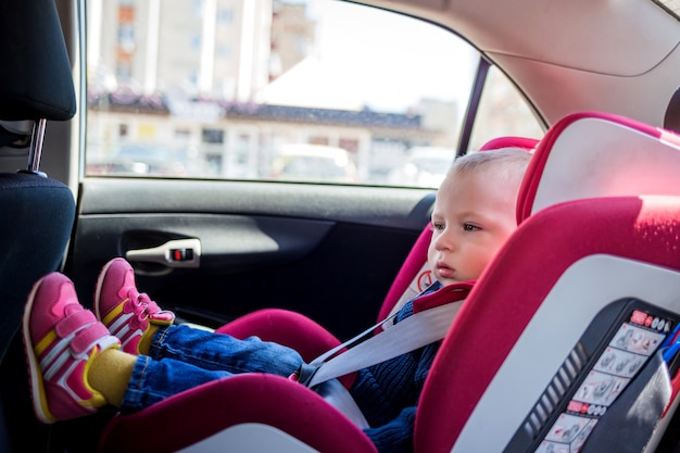 Ein kleiner Junge in einem roten Autositz. Ein kleines Baby in Jeans, Strickjacke und Turnschuhen sitzt auf einem Autositz. Sicherheit beim Transport von Kindern im Auto.