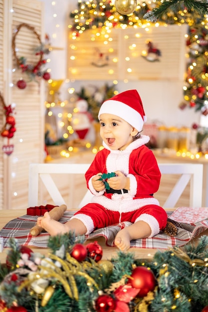 Ein kleiner Junge in einem Santa-Kostüm sitzt auf einem Tisch in der festlichen Neujahrsküche zu Hause, ein glückliches Kind feiert Weihnachten und Neujahr zu Hause