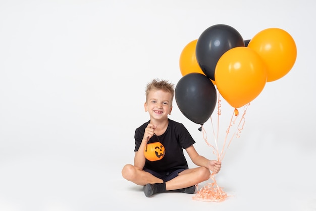 Ein kleiner Junge in einem schwarzen T-Shirt mit einem Korb für Leckereien und orangefarbenen und schwarzen Luftballons, die auf einem weißen Hintergrund sitzen