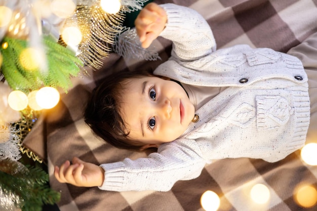 Ein kleiner Junge in einem Strickpullover liegt auf dem Bett vor dem Hintergrund eines festlich geschmückten Weihnachtsbaums zu Hause ein großes Porträt des Babys