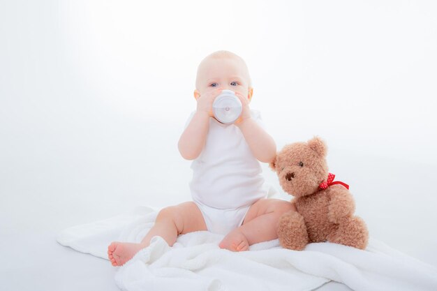 Ein kleiner Junge in einem weißen Body sitzt mit einem Teddybären auf weißem Hintergrund