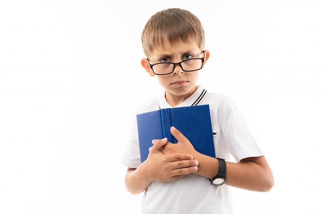 Ein kleiner Junge in einem weißen Hemd, blauen Shorts mit blonden Haaren, einer schwarzen Brille mit transparenter Brille, einem Armbanduhrständer und einem blauen Notizbuch ist im Dienst