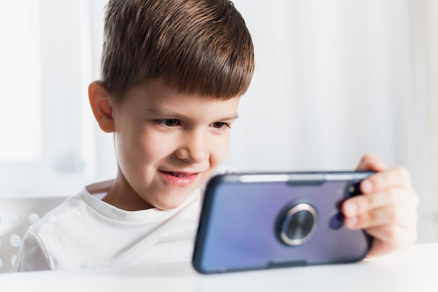 Ein kleiner Junge in einem weißen T-Shirt spielt zu Hause Spiele am Telefon. Ein glückliches Kind schaut auf sein Smartphone.