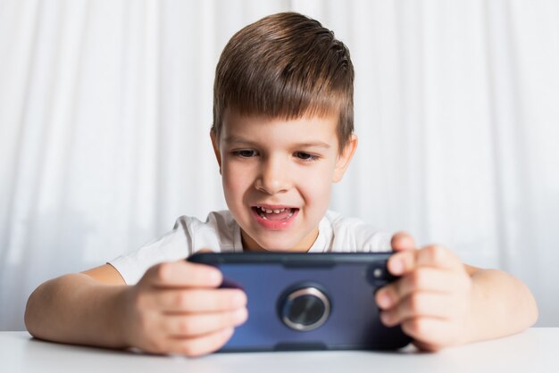 Ein kleiner Junge in einem weißen T-Shirt spielt zu Hause Spiele am Telefon. Ein glückliches Kind schaut auf sein Smartphone.