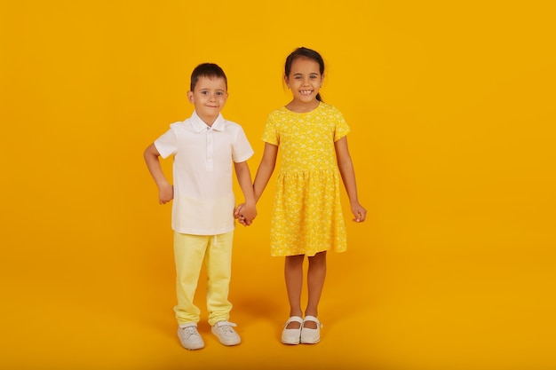 ein kleiner Junge in einem weißen T-Shirt und einer gelben Hose hält die Hand seiner Schwester in einem gelben Kleid