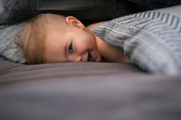 Ein kleiner Junge liegt auf dem Bauch im Bett und schaut in die Kamera und lacht.