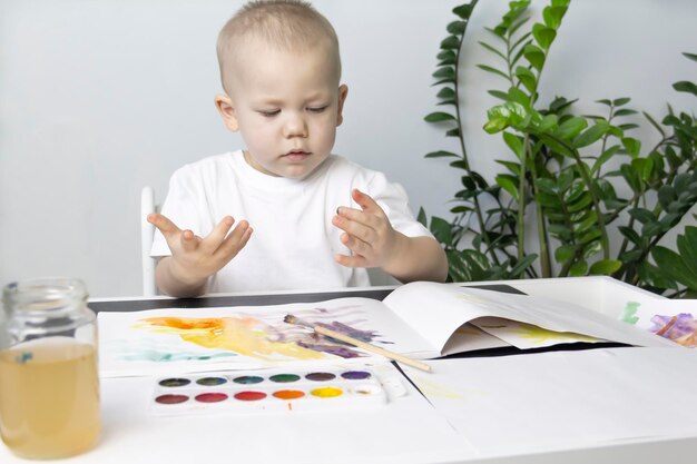 Ein kleiner Junge malt mit Aquarellfarben auf einem Tisch neben einer grünen Blume