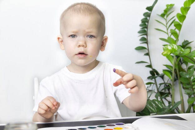 Ein kleiner Junge malt mit Aquarellfarben auf einem Tisch neben einer grünen Blume