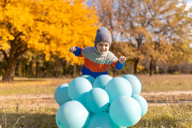 Ein kleiner Junge mit einem Armvoll Ballons geht im Herbstpark spazieren. Gelbe Bäume und blaue Kugeln. Stilvolles Kind