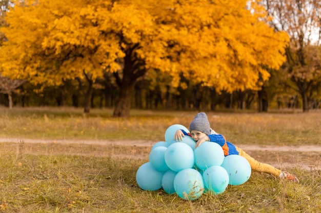 Ein kleiner Junge mit einem Armvoll Ballons geht im Herbstpark spazieren. Gelbe Bäume und blaue Kugeln. Stilvolles Kind.