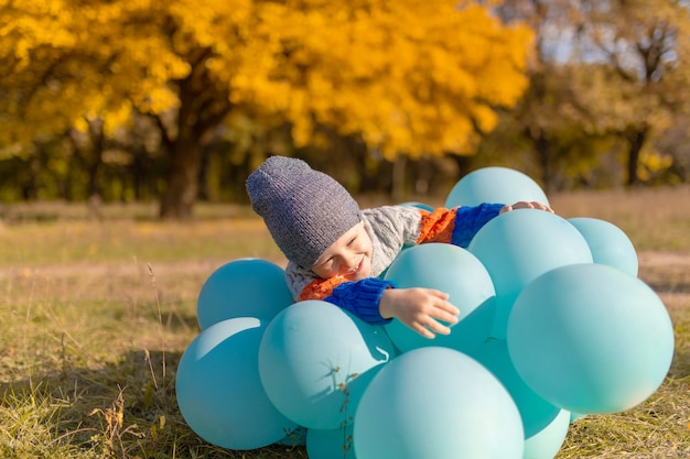 Ein kleiner Junge mit einem Armvoll Ballons geht im Herbstpark spazieren. Gelbe Bäume und blaue Kugeln. Stilvolles Kind.