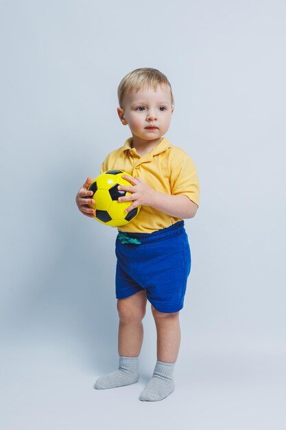 Ein kleiner Junge mit einem Fußball in seinen Händen auf einem weißen Hintergrund ein Kind ist ein Anfänger-Football-Spieler ein Sport für Kinder Kleiner Athlet Gelbe und blaue Fußball-Kit für Kinder